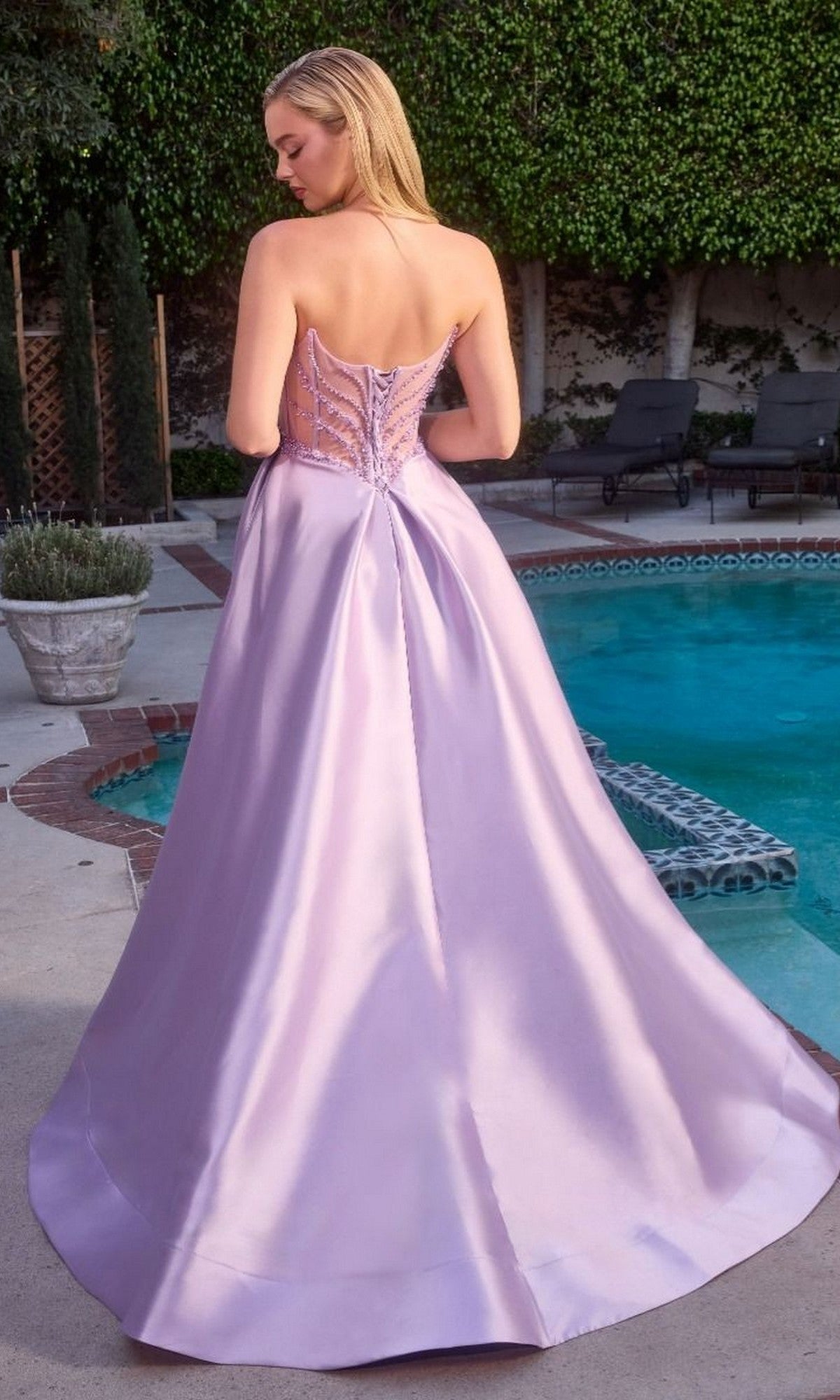 A woman wearing the Ladivine CD389 long prom dress with a strapless mikado bodice, a-line skirt, and lace-up back stands by a swimming pool outdoors.